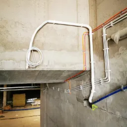 Instalacja rur i przewodów w surowym wnętrzu budynku, widoczne białe, szare, pomarańczowe i niebieskie rury przymocowane do betonowych ścian i sufitu, zwoje kabli zwisające z sufitu.