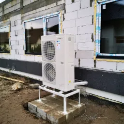 Biała pompa ciepła Innova zamontowana na metalowym stelażu przed budynkiem w trakcie budowy, widoczne okna z pianką montażową i niedokończona elewacja.