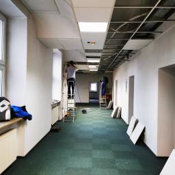 MTC Michał Hać - Montaż sufitu podwieszanego w długim korytarzu biurowym, dwaj pracownicy na drabinach, widoczne płyty sufitowe i narzędzia.