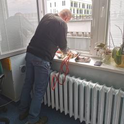 MTC Michał Hać - Mężczyzna w czarnej koszulce pochylony nad parapetem, prawdopodobnie w trakcie naprawy lub konserwacji urządzenia znajdującego się przy oknie, widoczne narzędzia, przewody, roślina doniczkowa...