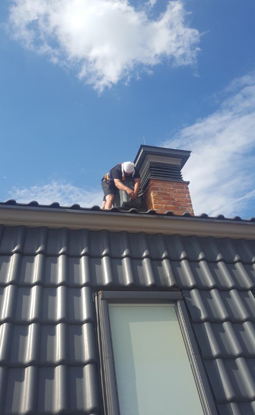 Pracownik w uprzęży zabezpieczającej, na dachu z dachówką ceramiczną, montuje element wentylacyjny na kominie z cegły klinkierowej, na tle błękitnego nieba.