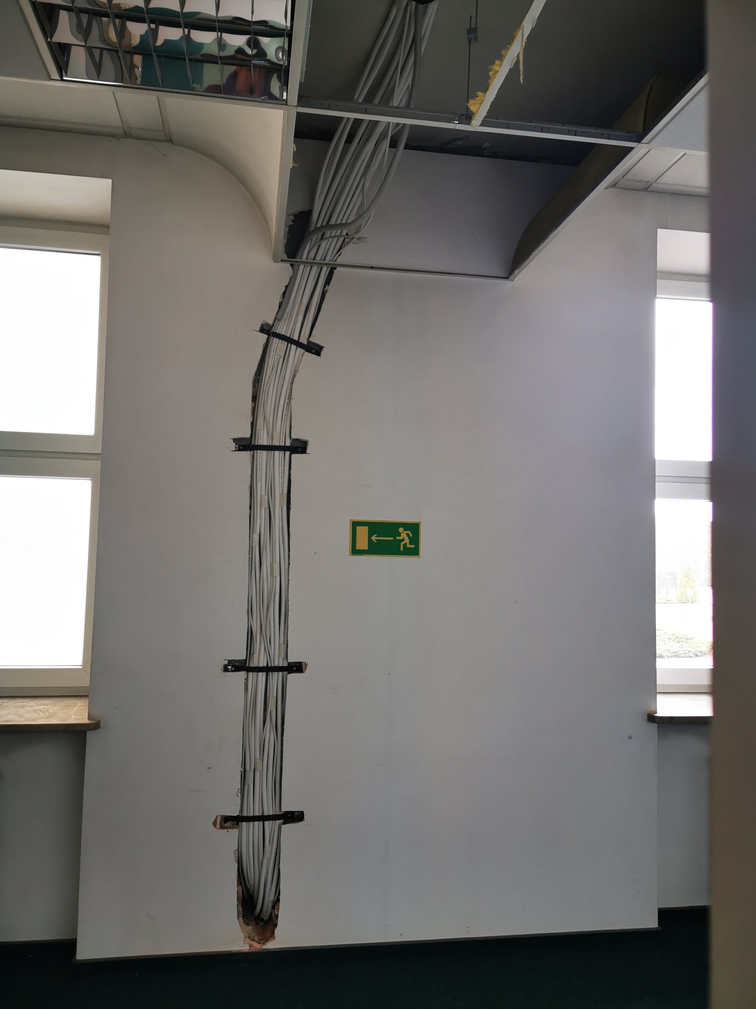 Pionowy kanał w ścianie biura z prowadzonymi kablami, zabezpieczonymi czarnymi uchwytami, widoczna instalacja podwieszanego sufitu i znak ewakuacyjny.