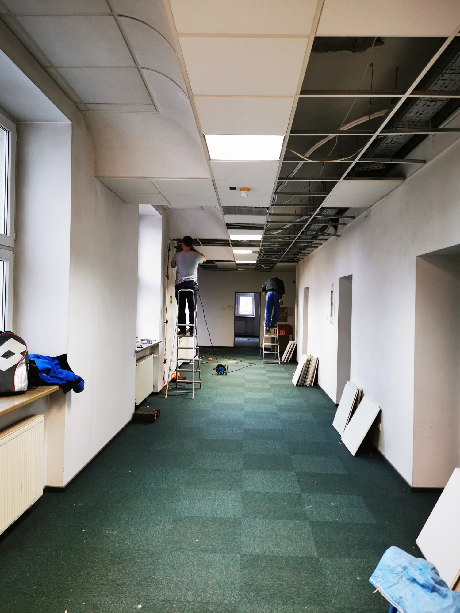 Montaż sufitu podwieszanego w długim korytarzu biurowym, dwaj pracownicy na drabinach, widoczne płyty sufitowe i narzędzia.