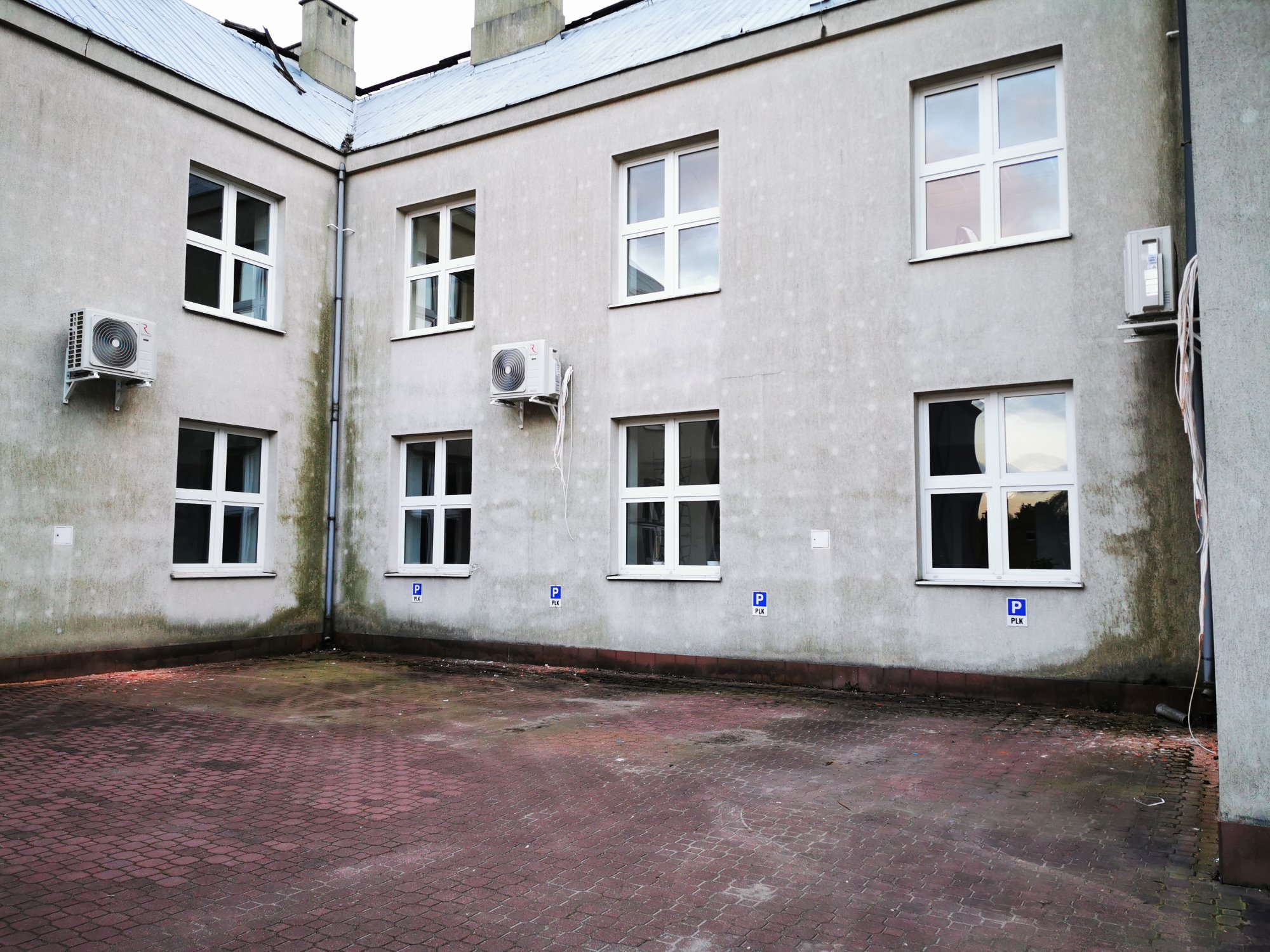Zewnętrzne jednostki klimatyzacji zamontowane na szarej ścianie budynku z licznymi oknami i parkingiem wyłożonym czerwoną kostką.