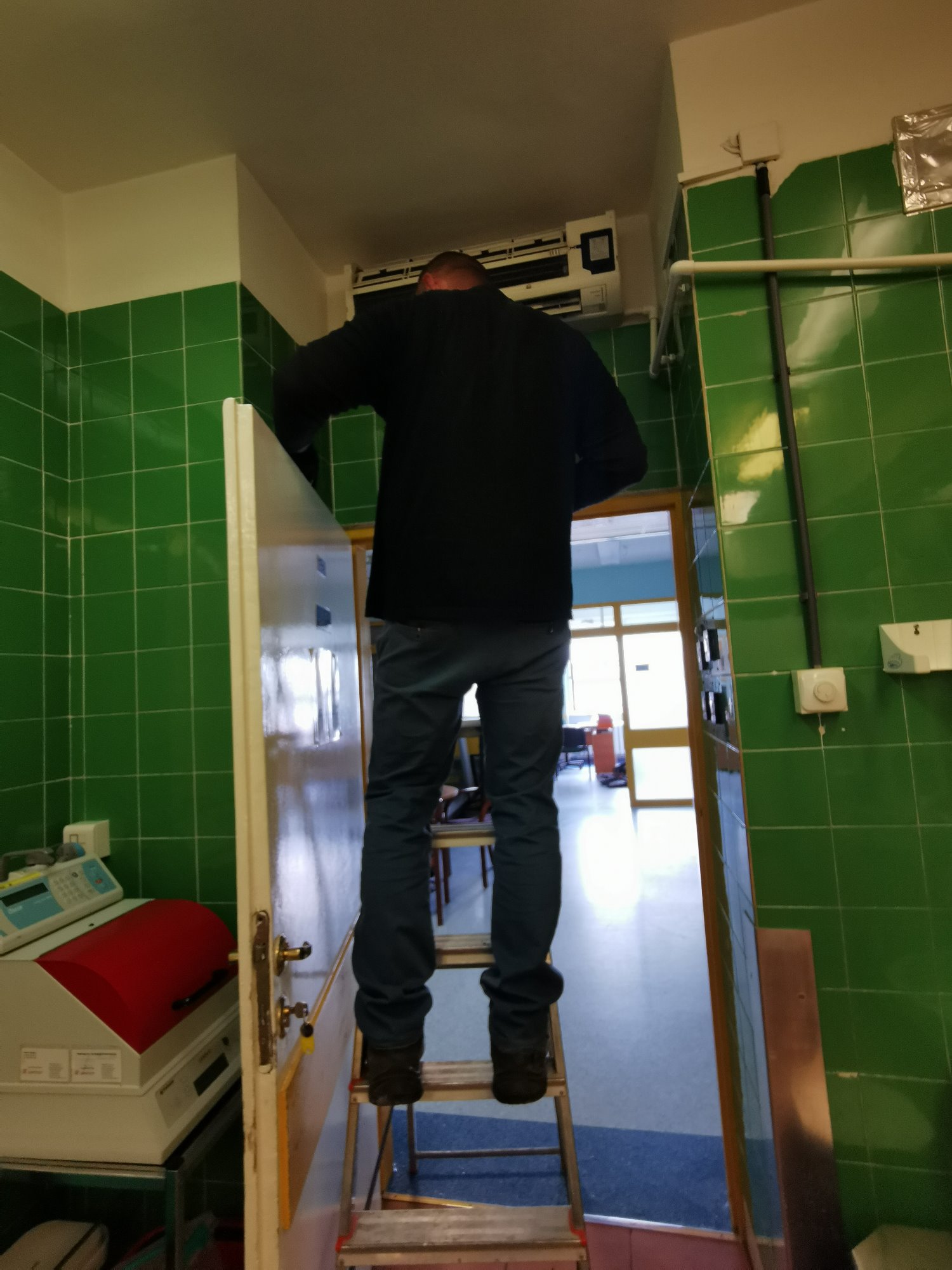 Instalator na drabinie w Józefowie, montujący klimatyzator w pomieszczeniu z zielonymi kafelkami na ścianach.