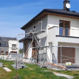 Dom jednorodzinny w trakcie prac elewacyjnych, widoczne rusztowania, częściowo otynkowana ściana, przygotowane materiały budowlane na trawniku.