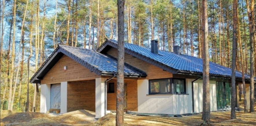 Nowoczesny dom jednorodzinny z drewnianą elewacją i ciemną dachówką, otoczony lasem sosnowym, z widocznym fragmentem ocieplonej ściany i przygotowanym terenem wokół budynku.