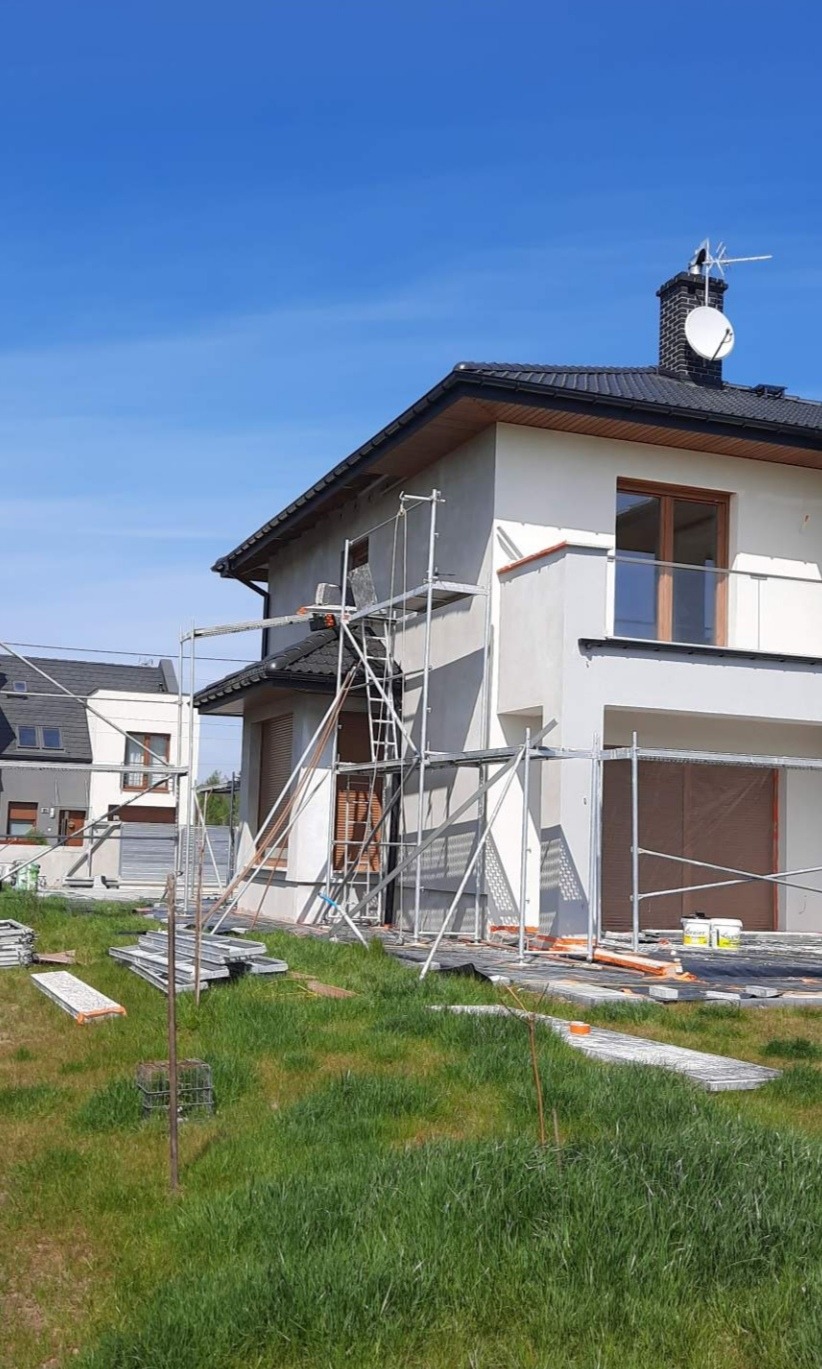 Dom jednorodzinny w trakcie prac elewacyjnych, widoczne rusztowania, częściowo otynkowana ściana, przygotowane materiały budowlane na trawniku.