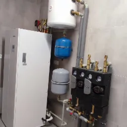 Biała pompa ciepła Kaisai z podłączonymi zbiornikami wyrównawczymi, rurami i rozdzielaczem, zamontowana na tle szarej, kafelkowanej ściany.