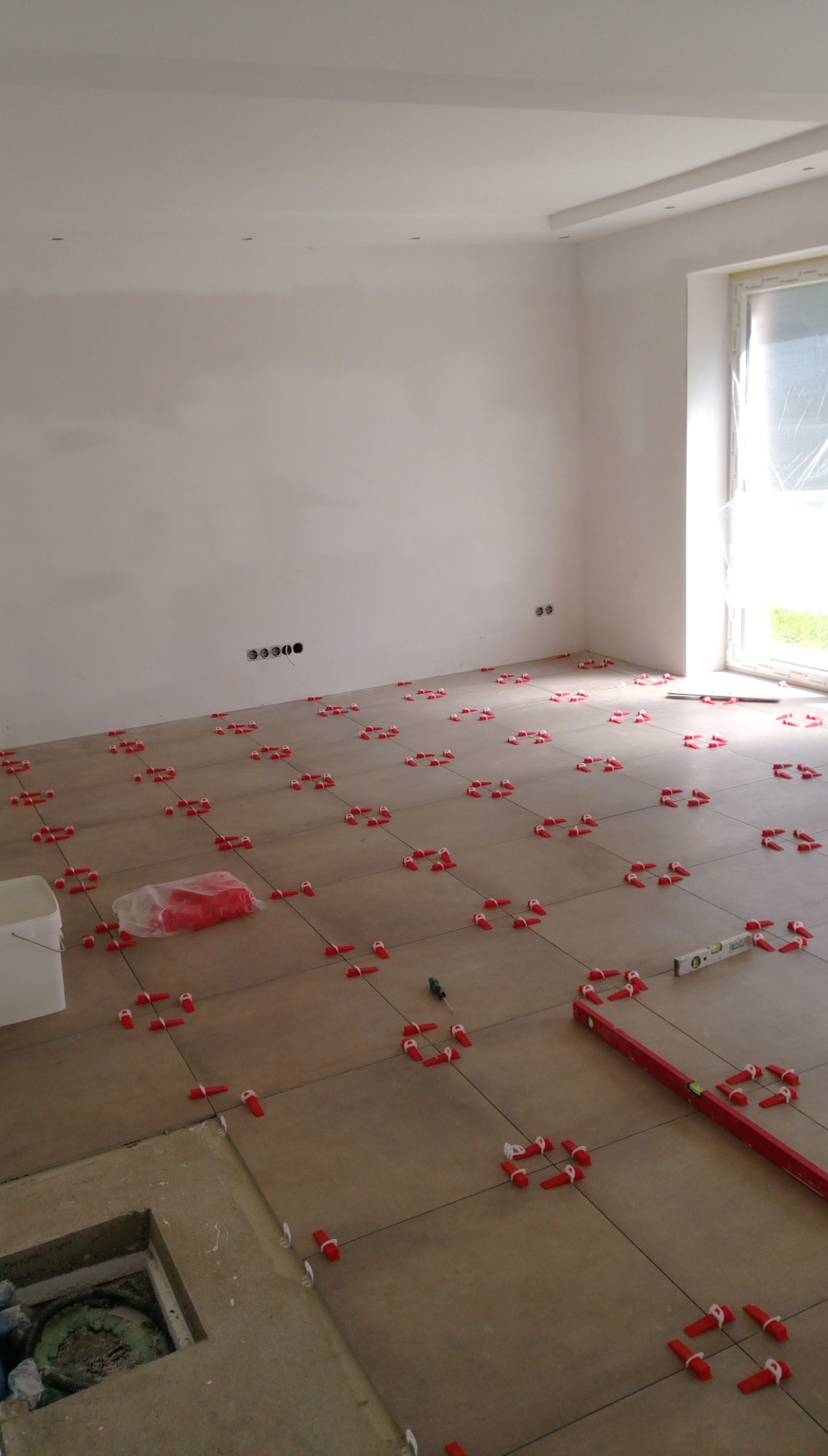 Układanie brązowych płytek podłogowych w jasnym pomieszczeniu, widoczne czerwone kliny i poziomica, w tle ściana z gniazdkami.