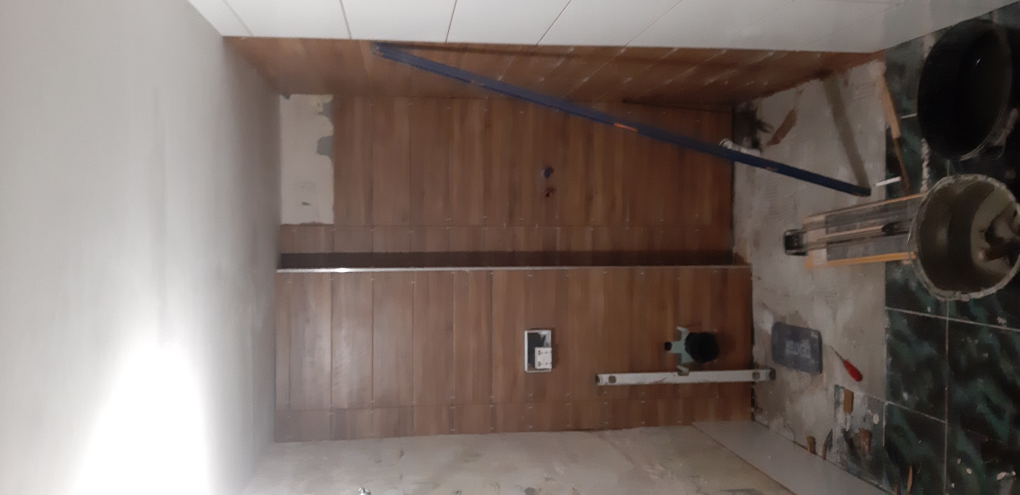 Wnętrze łazienki w trakcie remontu, widoczne drewnopodobne płytki na ścianach, poziomica, narzędzia glazurnicze i przygotowane podłoże pod posadzkę.