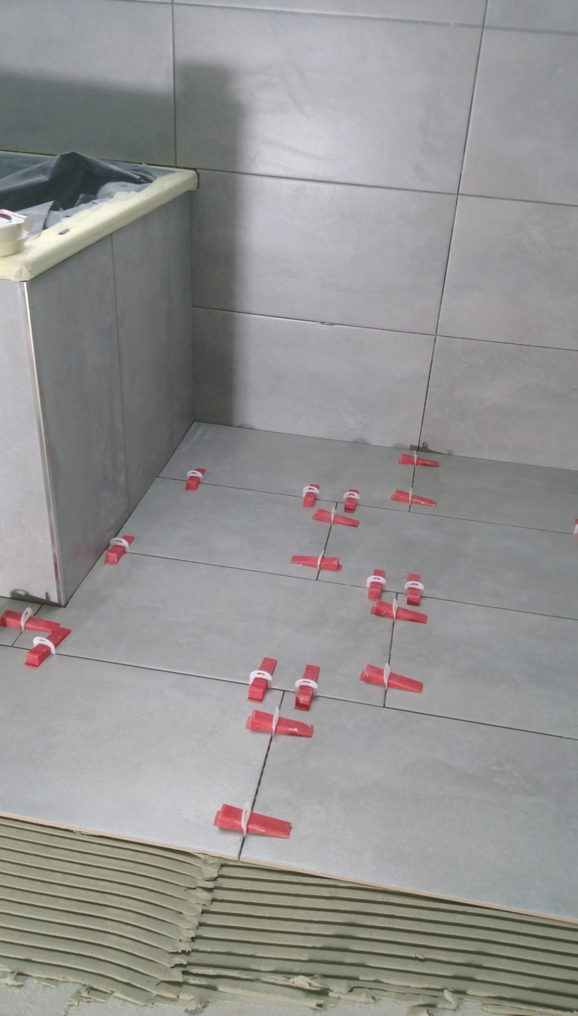 Szare płytki podłogowe w trakcie układania, z widocznymi czerwonymi klinami poziomującymi i warstwą kleju. Fragment zabudowy w tle.