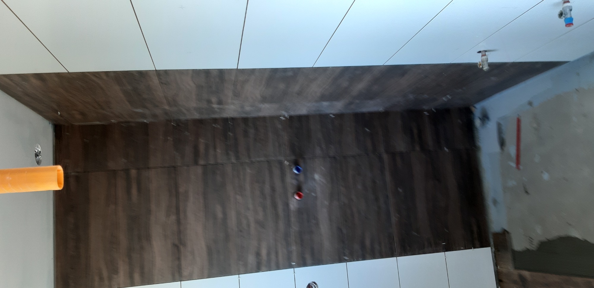 Ściana łazienki w trakcie wykańczania z brązowymi płytkami imitującymi drewno w strefie prysznica i białymi płytkami powyżej i poniżej, widoczne wystające rury instalacji wodnej i fragment...