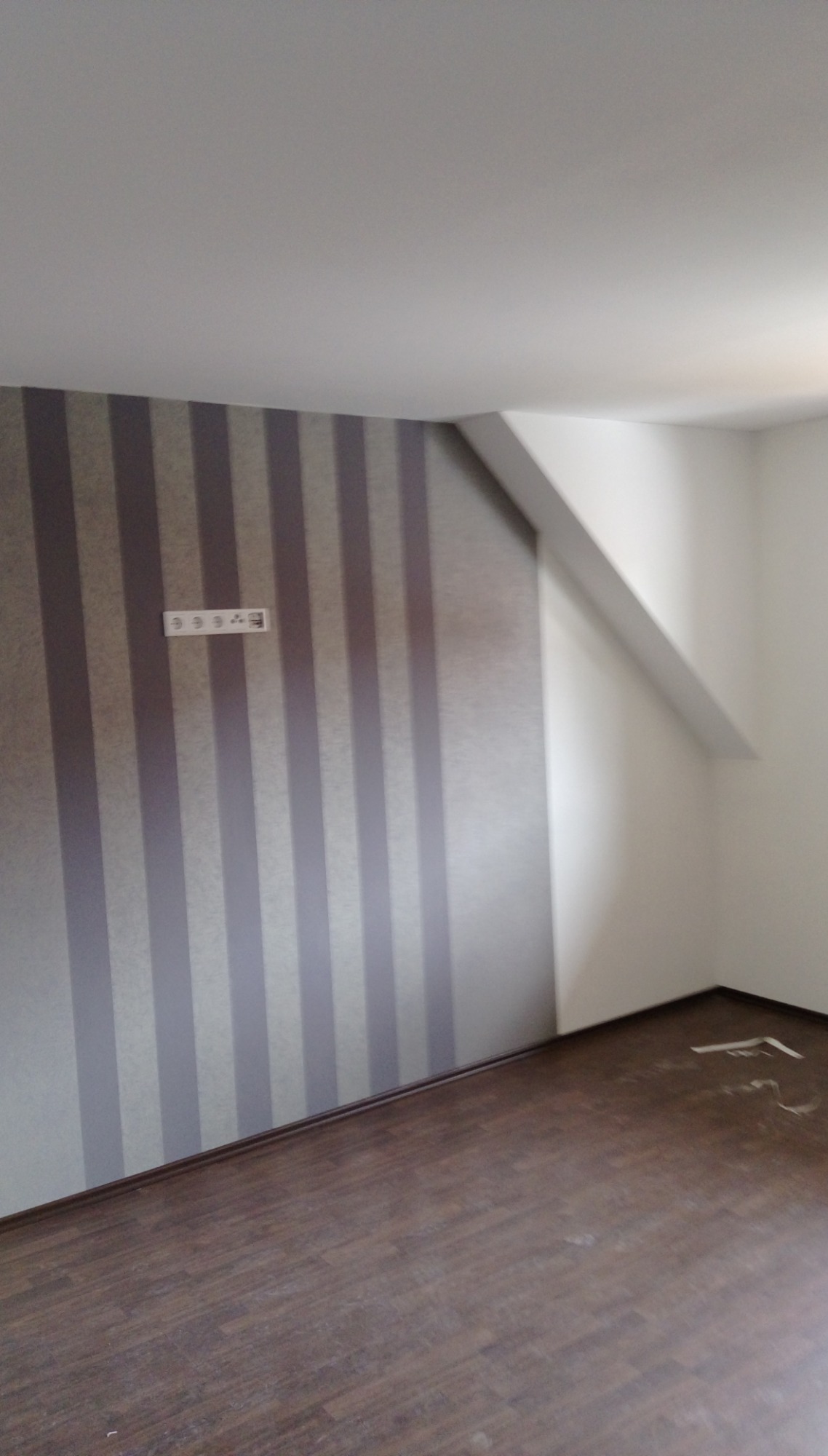 Pokój po remoncie z panelami podłogowymi w odcieniach brązu, ścianą w pionowe pasy szarości i białymi gładziami na pozostałych ścianach, widoczne gniazdka elektryczne.