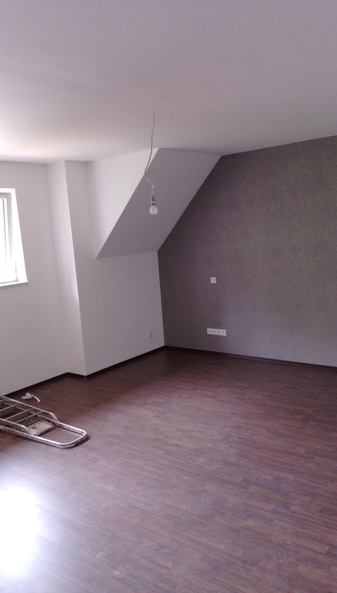 Pokój po remoncie: ciemne panele podłogowe, białe ściany z fragmentem pomalowanym na szaro, widoczna drabina malarska i wisząca żarówka bez klosza.