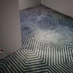 Wzorzysta wykładzina podłogowa z geometrycznym motywem w odcieniach szarości, bieli i zieleni, ułożona w jasnym pomieszczeniu z widocznymi przewodami elektrycznymi wystającymi ze ściany.