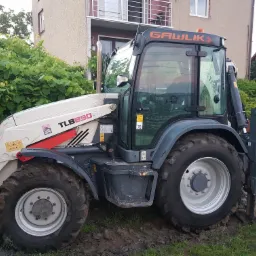 Ciężka maszyna budowlana TLB890 marki Terex z napisem GAWLIK na tle budynku i zieleni, widoczne ślady błota na oponach.