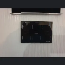 Czarna, szklana płyta indukcyjna marki DEIMIC zamontowana na białej ścianie z delikatnym wzorem, widoczny fragment zabudowy kuchennej nad płytą.