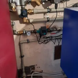 Złożona instalacja hydrauliczna i grzewcza z licznymi zaworami, pompami obiegowymi i manometrami, zamontowana na ścianie o nierównym wykończeniu, widoczny fragment czerwonego zbiornika...