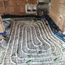 Rozłożone białe rury ogrzewania podłogowego na folii z siatką, widoczne kolektory i nieotynkowane ściany z cegły dziurawki w tle.