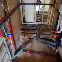 Wnętrze łazienki w trakcie remontu, widoczne kolorowe rury instalacji wodnej i kanalizacyjnej, zamontowana podtynkowa spłuczka Grohe, surowe drewniane ściany i podłoga w trakcie prac hydraulicznych.