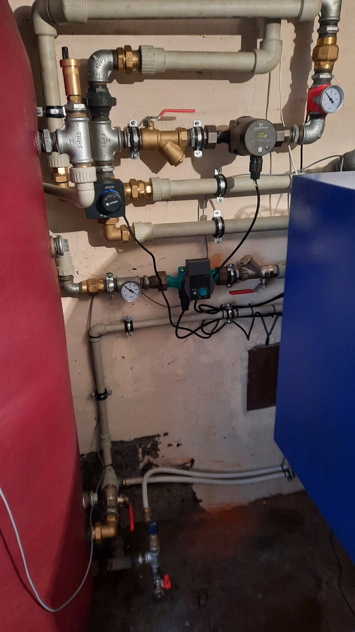 Złożona instalacja hydrauliczna i grzewcza z licznymi zaworami, pompami obiegowymi i manometrami, zamontowana na ścianie o nierównym wykończeniu, widoczny fragment czerwonego zbiornika...