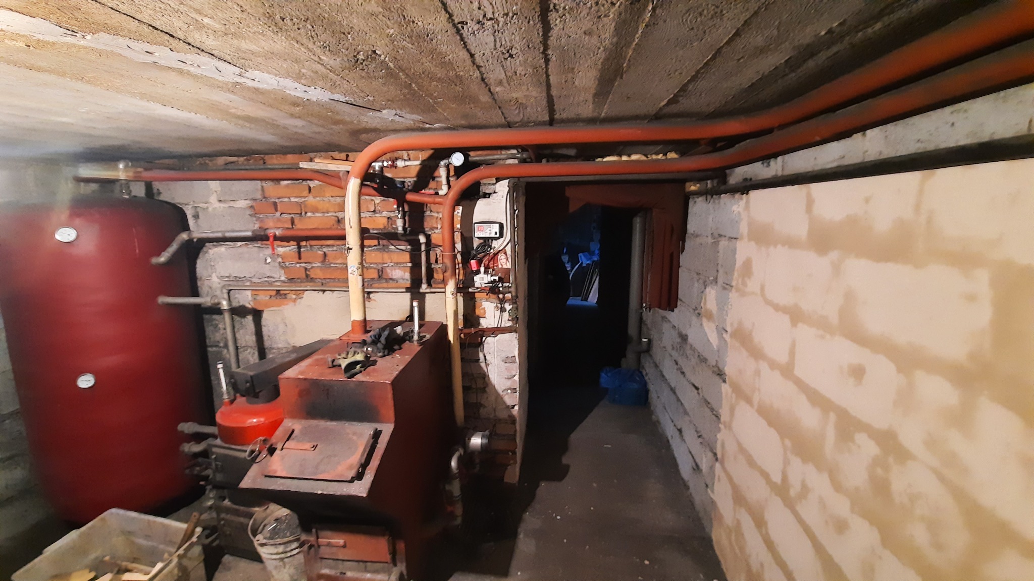 Piwnica z instalacją centralnego ogrzewania: czerwony zbiornik akumulacyjny, piec na paliwo stałe, rury miedziane i sterownik na ścianie z cegły.