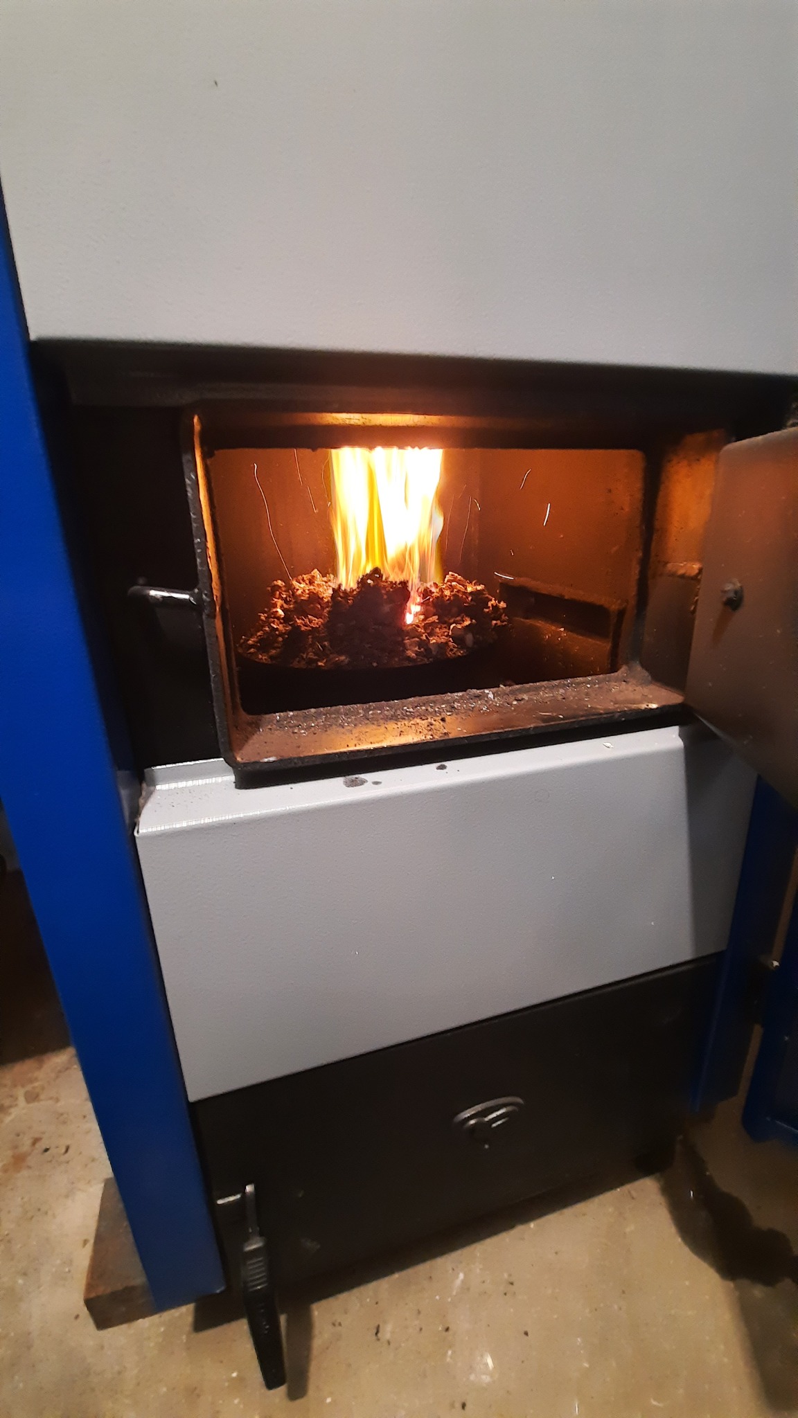 Otwarty piec centralnego ogrzewania z palącym się węglem, widoczne płomienie i żarzące się bryły paliwa, oświetlające wnętrze paleniska.