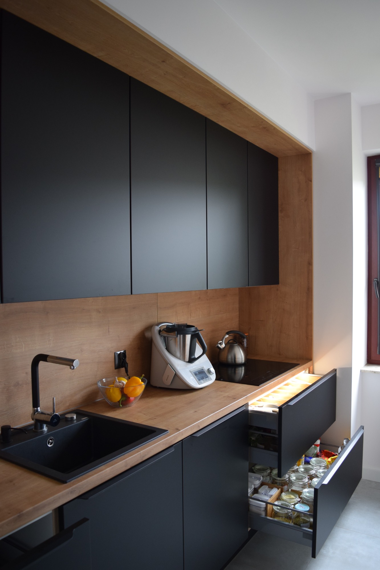 Nowoczesna kuchnia z czarnymi szafkami i drewnianym blatem, otwarta szuflada z zapasami, na blacie czajnik, Thermomix i miska z owocami, widoczny zlew z czarną baterią.