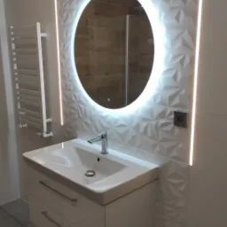 Nowoczesna łazienka z umywalką nablatową, szafką z szufladami, okrągłym lustrem z podświetleniem LED i grzejnikiem drabinkowym na ścianie z trójwymiarowymi płytkami.