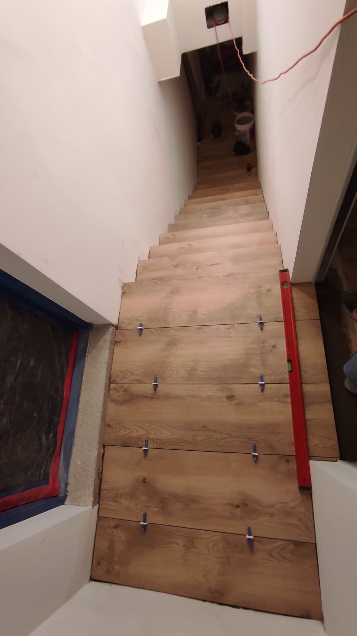 Układanie płytek imitujących drewno na schodach, widoczny poziomica i krzyżyki dystansowe, surowy stan wykończenia ścian.