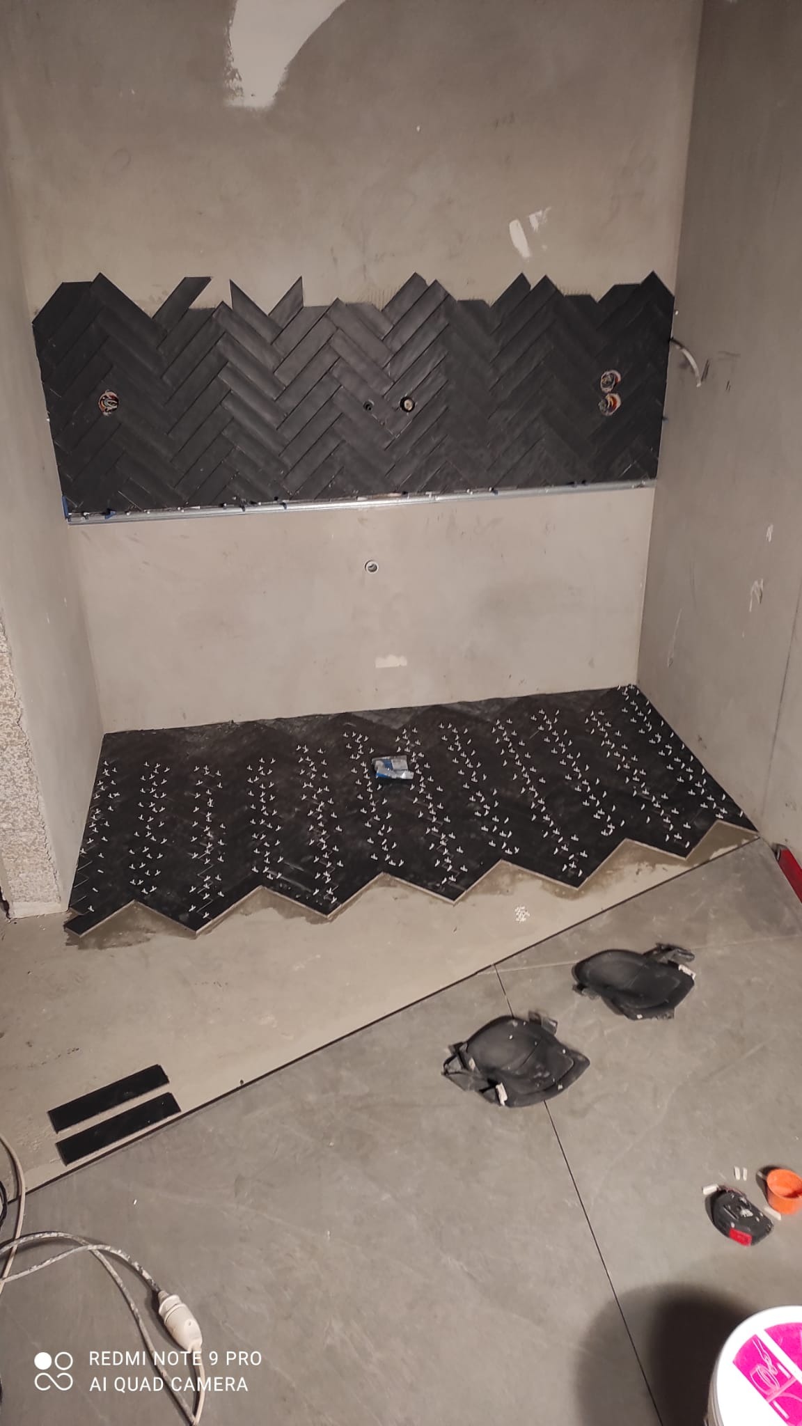 Montaż ciemnych, cegiełkowych płytek w jodełkę na ścianie i podłodze w surowej łazience, widoczne krzyżyki dystansowe i nakolanniki.