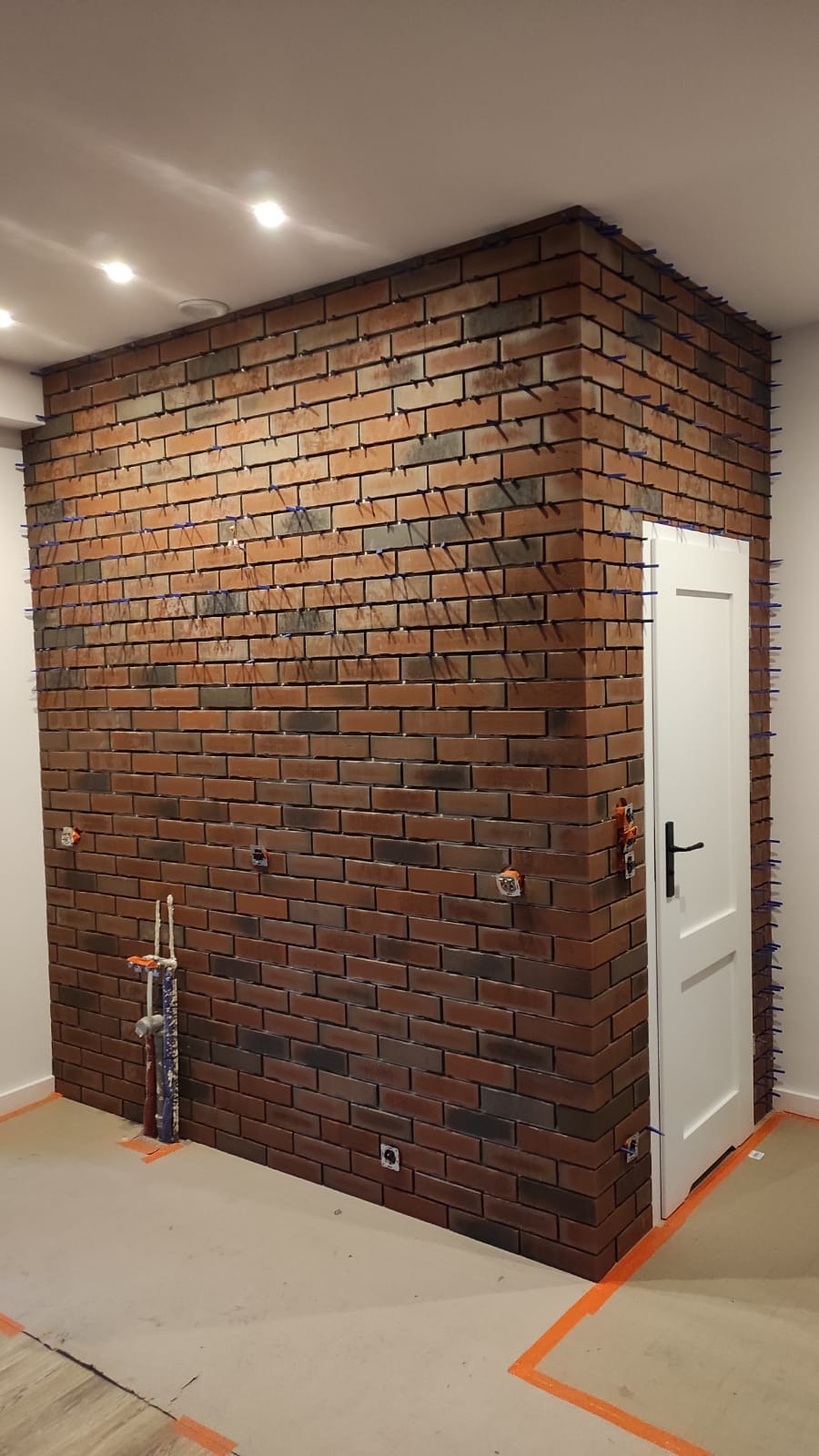 Ściana wyłożona cegłą klinkierową w odcieniach czerwieni i brązu, z widocznymi niebieskimi klinami dystansowymi, obok białe drzwi z czarną klamką, instalacja elektryczna wystająca ze ściany...