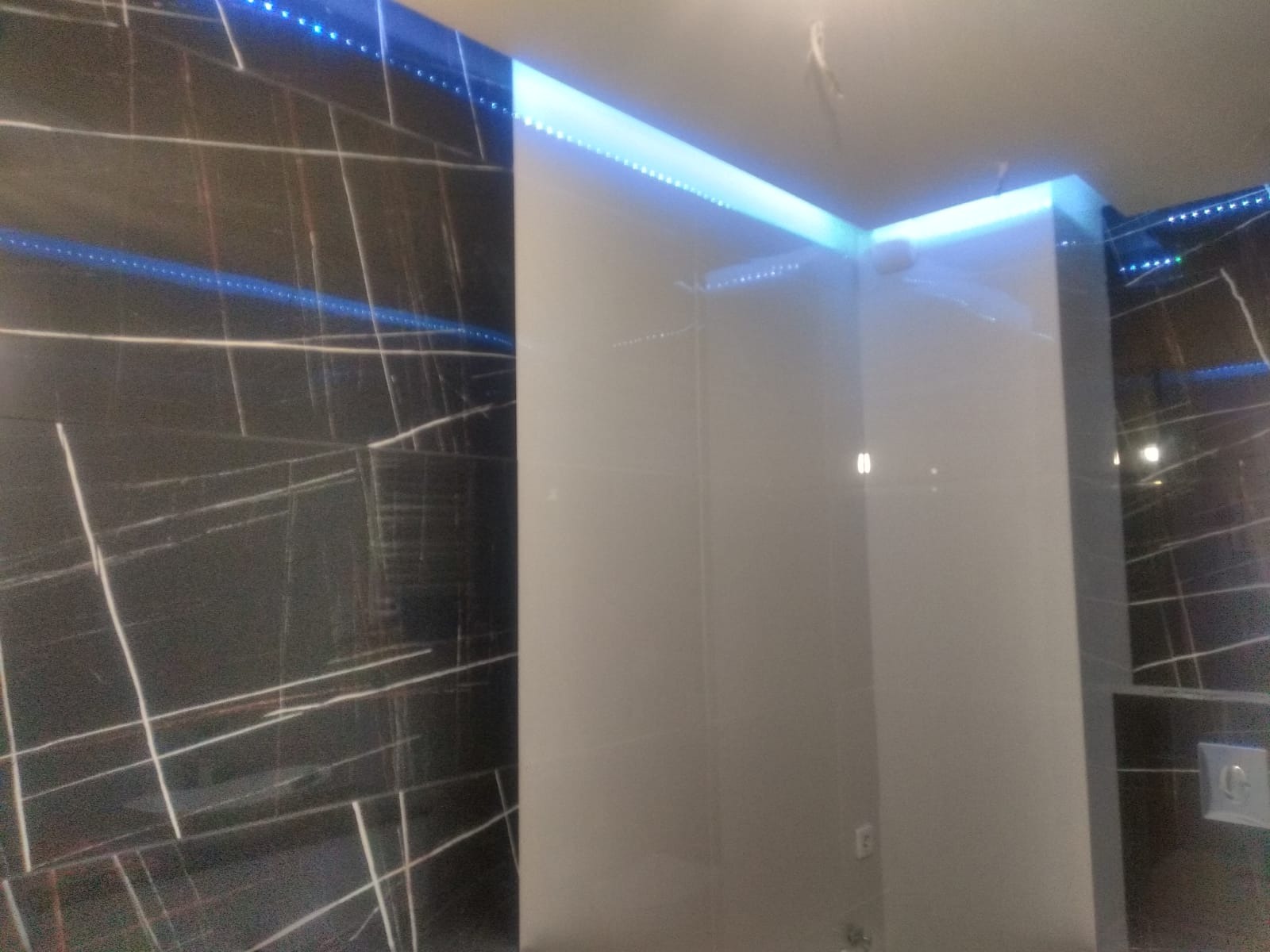 Nowoczesna łazienka z czarną marmurową ścianą akcentową i podświetleniem LED wzdłuż sufitu, tworzącym niebieską poświatę.