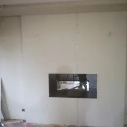 Ściana pokryta płytami gipsowo-kartonowymi z wyciętym otworem, widoczne ślady po wkrętach, na podłodze płytki w stylu rustykalnym, narzędzia i resztki materiałów budowlanych.