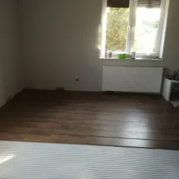 Układanie paneli podłogowych w pokoju z brązowymi panelami, widoczne białe podkłady izolacyjne i opakowania paneli, okno z roletami i grzejnikiem.