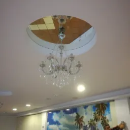 Kryształowy żyrandol zwisający z okrągłego otworu w suficie, w którym widać kafelki, a mężczyzna w czerwonej koszuli w kratę montuje coś na ścianie z fototapetą z motywem plaży.