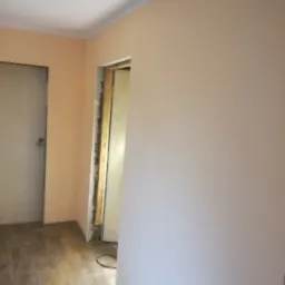 Świeżo pomalowane ściany w odcieniach brzoskwini w pokoju z drewnianą podłogą, widoczne ościeżnice drzwiowe w trakcie montażu.