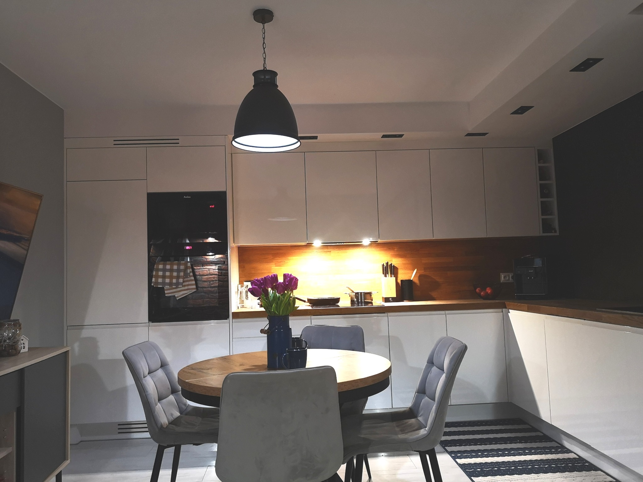 Nowoczesna kuchnia z białymi szafkami, drewnianym blatem, okrągłym stołem i szarymi krzesłami tapicerowanymi, oświetlona wiszącą lampą w stylu industrialnym.