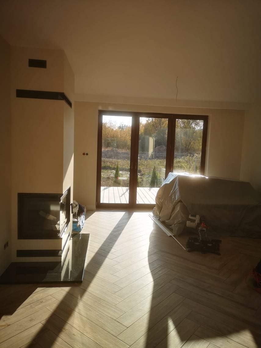 Salon w trakcie wykańczania: parkiet ułożony w jodełkę, kominek z czarnym obramowaniem, duże okno tarasowe z widokiem na ogród, meble zakryte folią ochronną, wpadające promienie słoneczne.