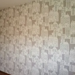 Ściana pokryta szarobrązową tapetą z rysunkami białych domk&oacute;w i drzew, tworząca wz&oacute;r przypominający miasteczko.