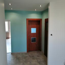 Hadson - Korytarz po remoncie z dwiema drewnianymi drzwiami z przeszkleniami, pomalowany na biało i turkusowo, z szarą podłogą w płytkach i punktowym oświetleniem w suficie.