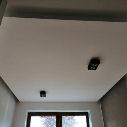 Hadson - Pomieszczenie po malowaniu sufitu na jasny kolor z zamontowanymi dwoma czarnymi lampami punktowymi. Ściany częściowo przygotowane do dalszych prac wykończeniowych, widoczne ślady po szpachlowaniu.