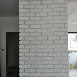 Hadson - Fragment ściany wykończonej białymi płytkami imitującymi cegłę, widoczny w trakcie remontu wnętrza, z fragmentem niezakończonej gładzi na suficie i oknem w tle.