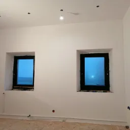 Wnętrze pokoju po malowaniu ścian na biało, widoczne dwa okna z czarnymi ramami, instalacja elektryczna w ścianach i rury grzewcze przy podłodze, surowa podłoga przygotowana do dalszych prac...