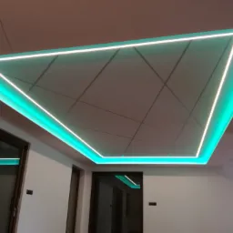 Podwieszany sufit z płyt z podświetleniem LED w kształcie trójkąta, widoczne linie podziału płyt i wisząca lampa z kablem.