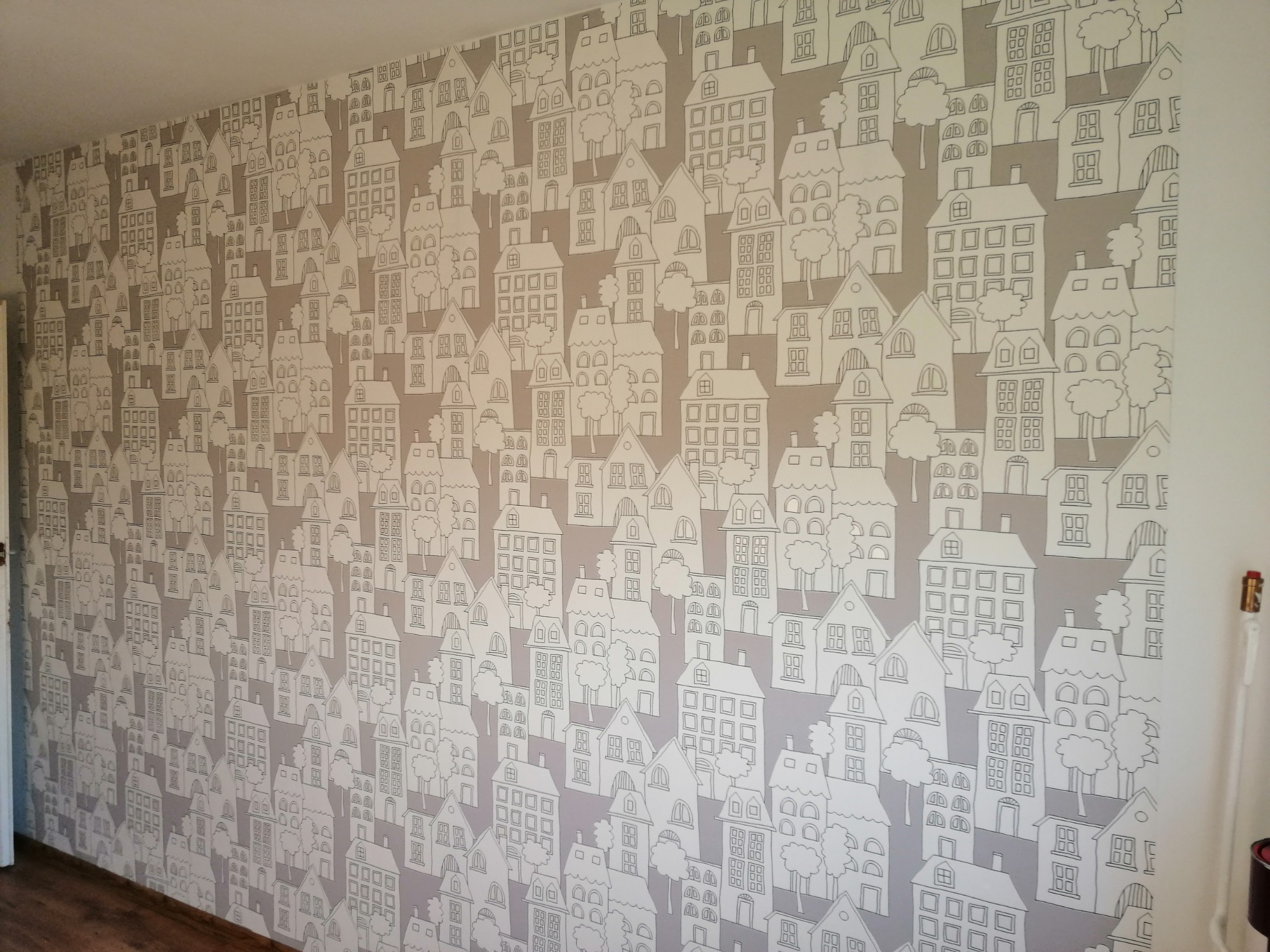 Ściana pokryta szarobrązową tapetą z rysunkami białych domków i drzew, tworząca wzór przypominający miasteczko.