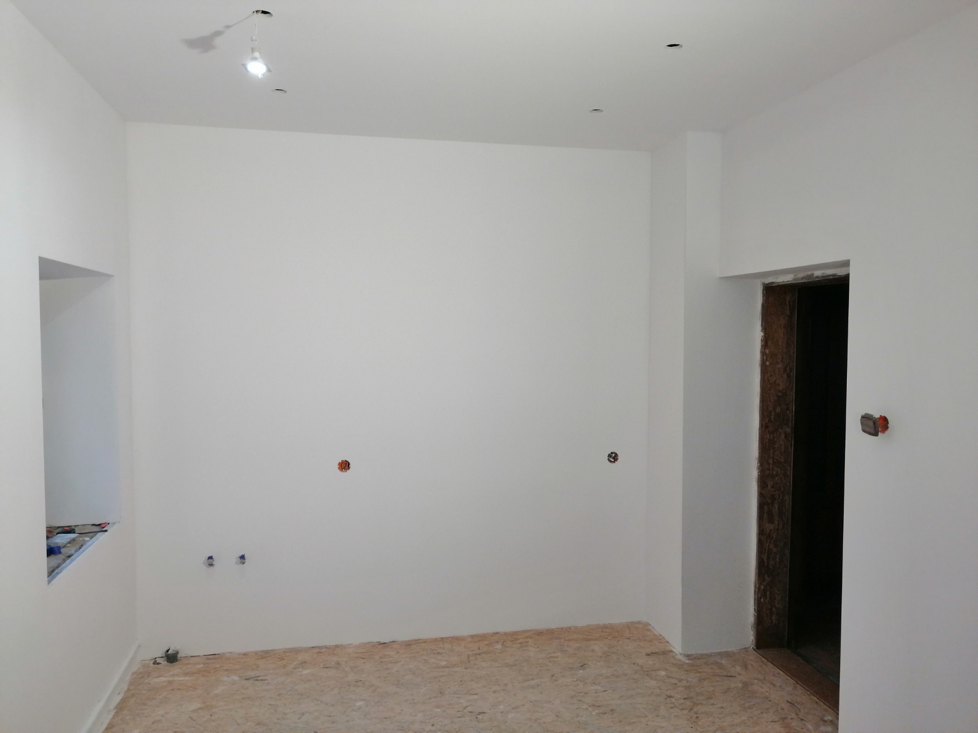 Wnętrze pokoju po świeżym malowaniu na biało, z widocznymi puszkami elektrycznymi i przygotowanym otworem okiennym, podłoga wyłożona płytami OSB.