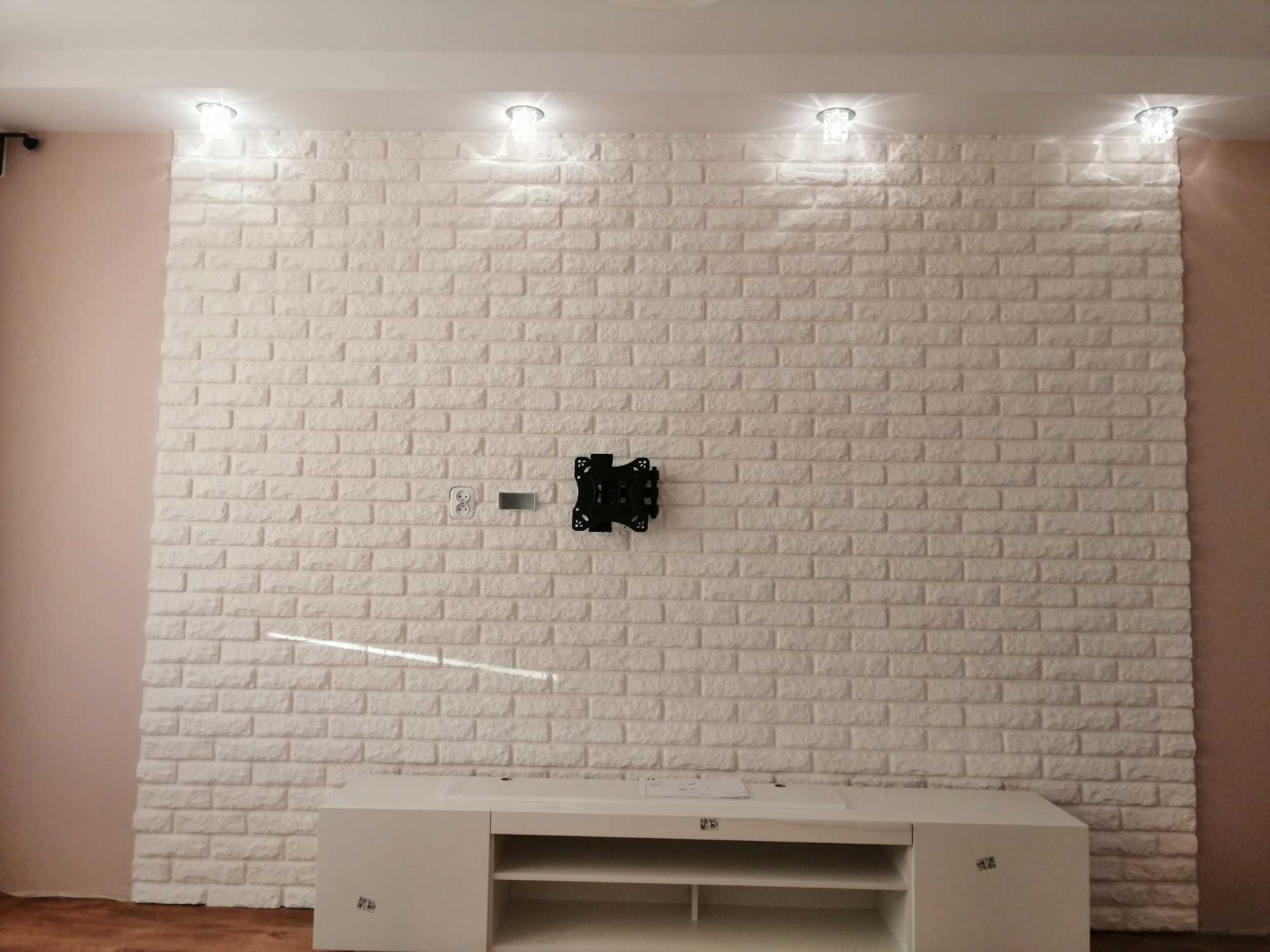 Ściana wykończona białą cegłą dekoracyjną z zamontowanym uchwytem do telewizora, poniżej biała szafka RTV i cztery punktowe lampy sufitowe.