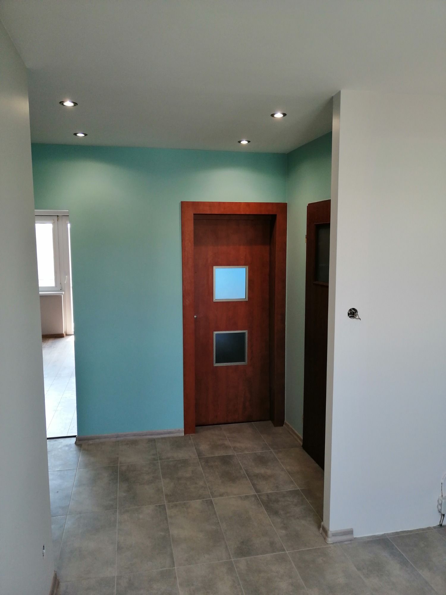 Korytarz po remoncie z dwiema drewnianymi drzwiami z przeszkleniami, pomalowany na biało i turkusowo, z szarą podłogą w płytkach i punktowym oświetleniem w suficie.